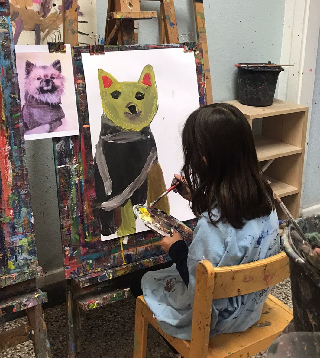 MiK Jugendkunstschule Berlin Mitte - Das Foto zeigt ein junges Mädchen mit braunen Haaren, das vor einer Staffelei sitzt und einen Hund malt, anhand eines daneben hängenden Fotos